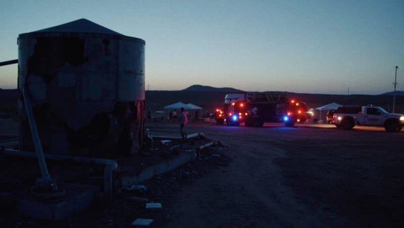 Tanque de almacenamiento de agua dañado en zona desértica con pipas de emergencia y luces de trabajo al atardecer