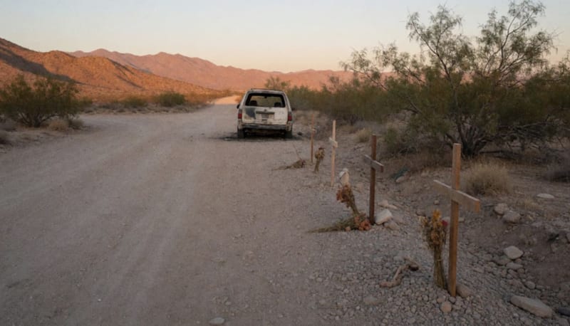 Camioneta calcinada junto a un camino de terracería en el desierto con cruces de madera y montañas al fondo al atardecer