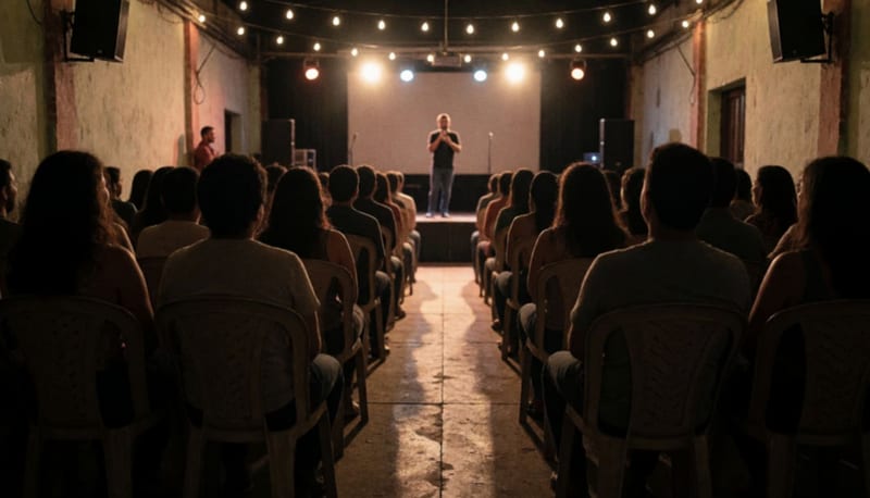 Público riendo sentado en sillas de plástico frente a un pequeño escenario iluminado donde un comediante sostiene un micrófono