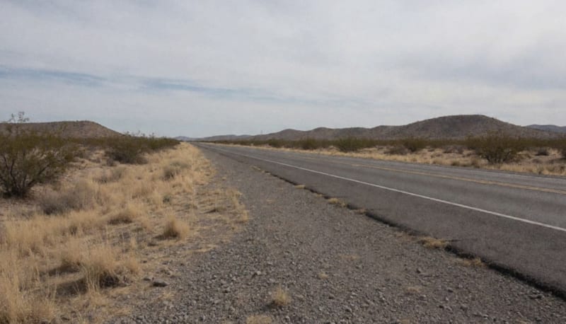 Tramo despejado de una carretera rural entre matorral desértico, visto desde la orilla del camino