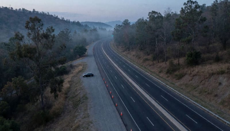 Vista aérea de una carretera sinuosa entre cerros boscosos al anochecer, con un automóvil oscuro detenido en el acotamiento