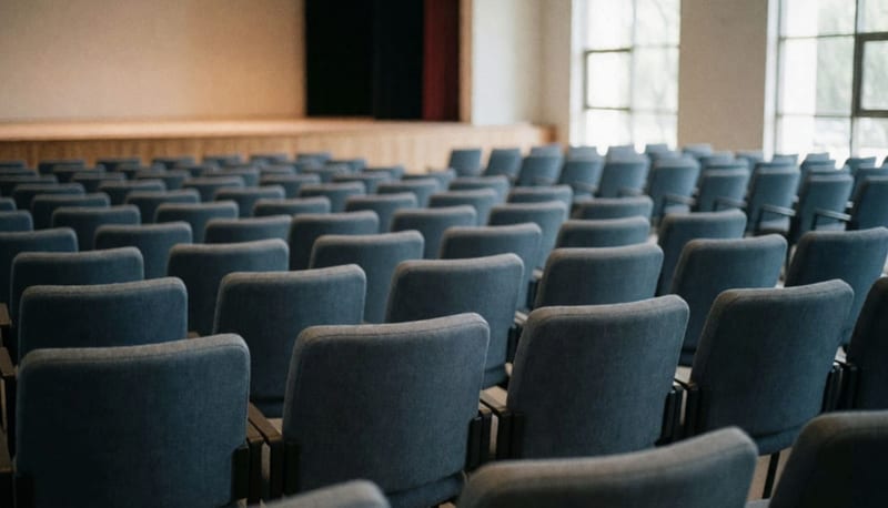 Sala de conferencias con filas de asientos vacíos frente a un escenario, luz natural por las ventanas