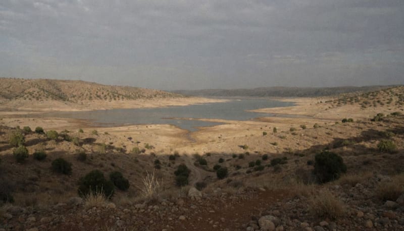 Una presa en región árida con agua reflejando un cielo nublado y cerros secos alrededor