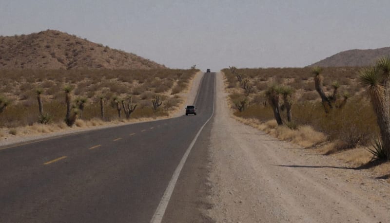 Carretera recta de dos carriles en zona desértica con una camioneta oscura a lo lejos