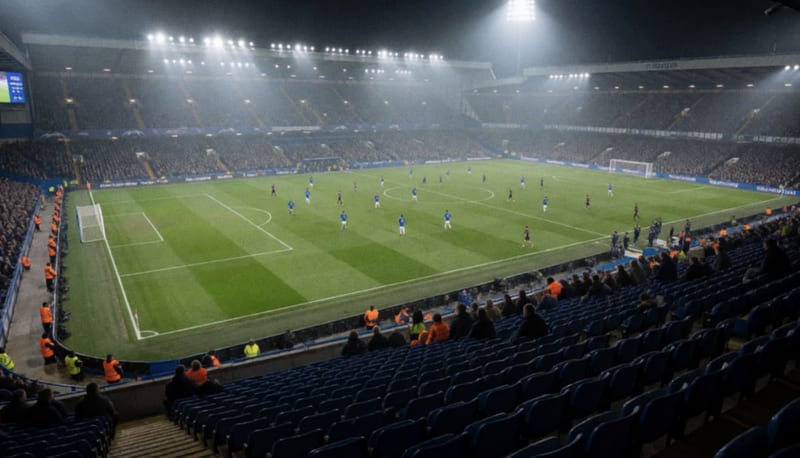 Estadio de futbol de noche visto desde las gradas, con jugadores diminutos sobre el campo iluminado por reflectores y asientos vacíos al frente