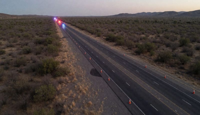 Vista aérea de una carretera entre matorrales secos al anochecer, con luces rojas y azules a lo lejos sobre el asfalto