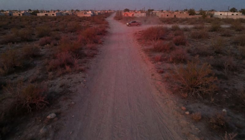 Brecha de terracería entre matorrales secos al atardecer, con una camioneta a lo lejos y luces rojas y azules sobre el polvo