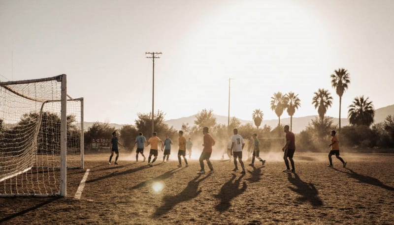 Cancha de fútbol en polvo con jugadores preparándose para el partido, cercada por vegetación desértica