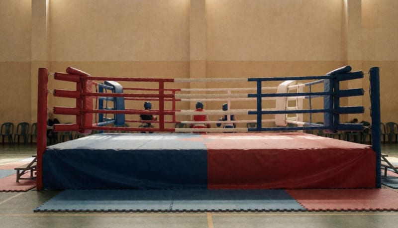 Ring de combate con esquinas acolchadas en un gimnasio amplio, con tres pequeñas figuras de espaldas junto a las cuerdas