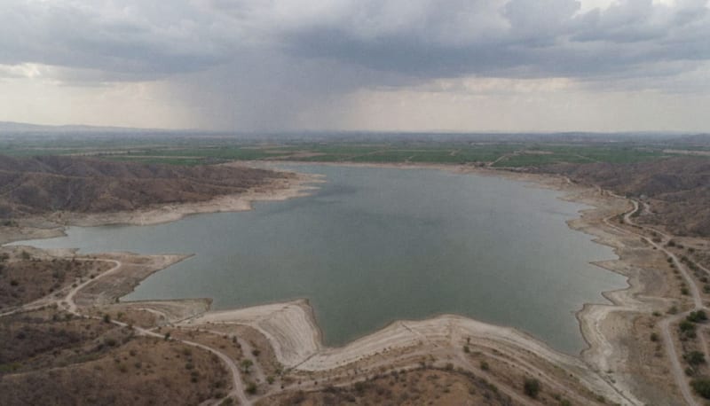 Vista aérea de un embalse en el desierto con orillas secas expuestas, cerros áridos y nubes de lluvia al fondo