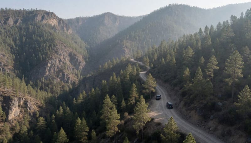 Vista aérea de un camino de terracería entre cañones y bosques de pino en la sierra, con dos camionetas oscuras a lo lejos