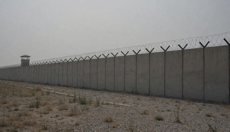 Muro de concreto con alambre de navajas y una torre de vigilancia al fondo bajo cielo nublado