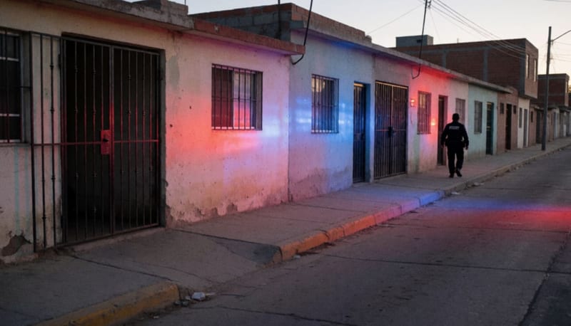 Calle residencial de madrugada con luces rojas y azules sobre las casas y un policía a lo lejos visto de espaldas
