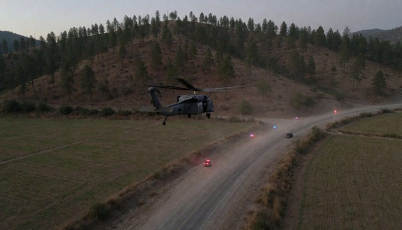 Helicóptero sobrevolando una carretera rural entre campos y cerros boscosos al atardecer, con luces rojas y azules a lo lejos