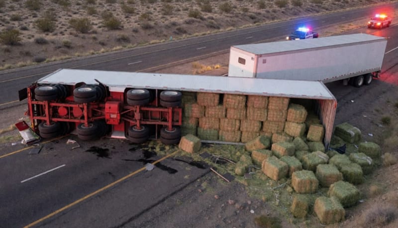 Tráiler volcado sobre la carretera con fardos de alfalfa dispersos en el pavimento y el desierto de fondo